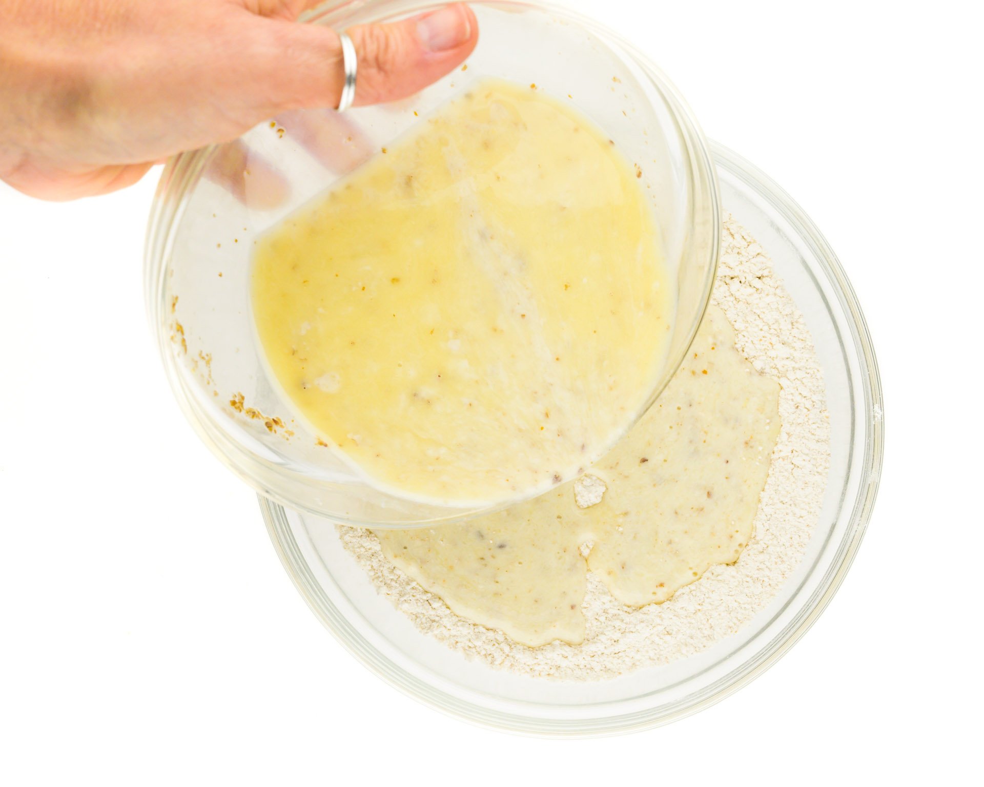 A hand holds a bowl of liquid ingredients and is pouring it into a bowl of flour ingredients.
