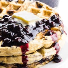 A stack of blueberry waffles has blueberry sauce on top with a pat of vegan butter.