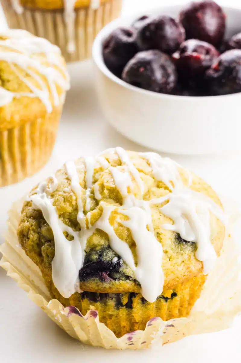 A muffin has lots of frosting drizzled on top with more muffins behind it and a bowl of frozen cherries.
