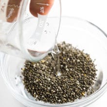 A hand holds a glass measuring cup with water and is pouring it into a bowl with chia seeds.