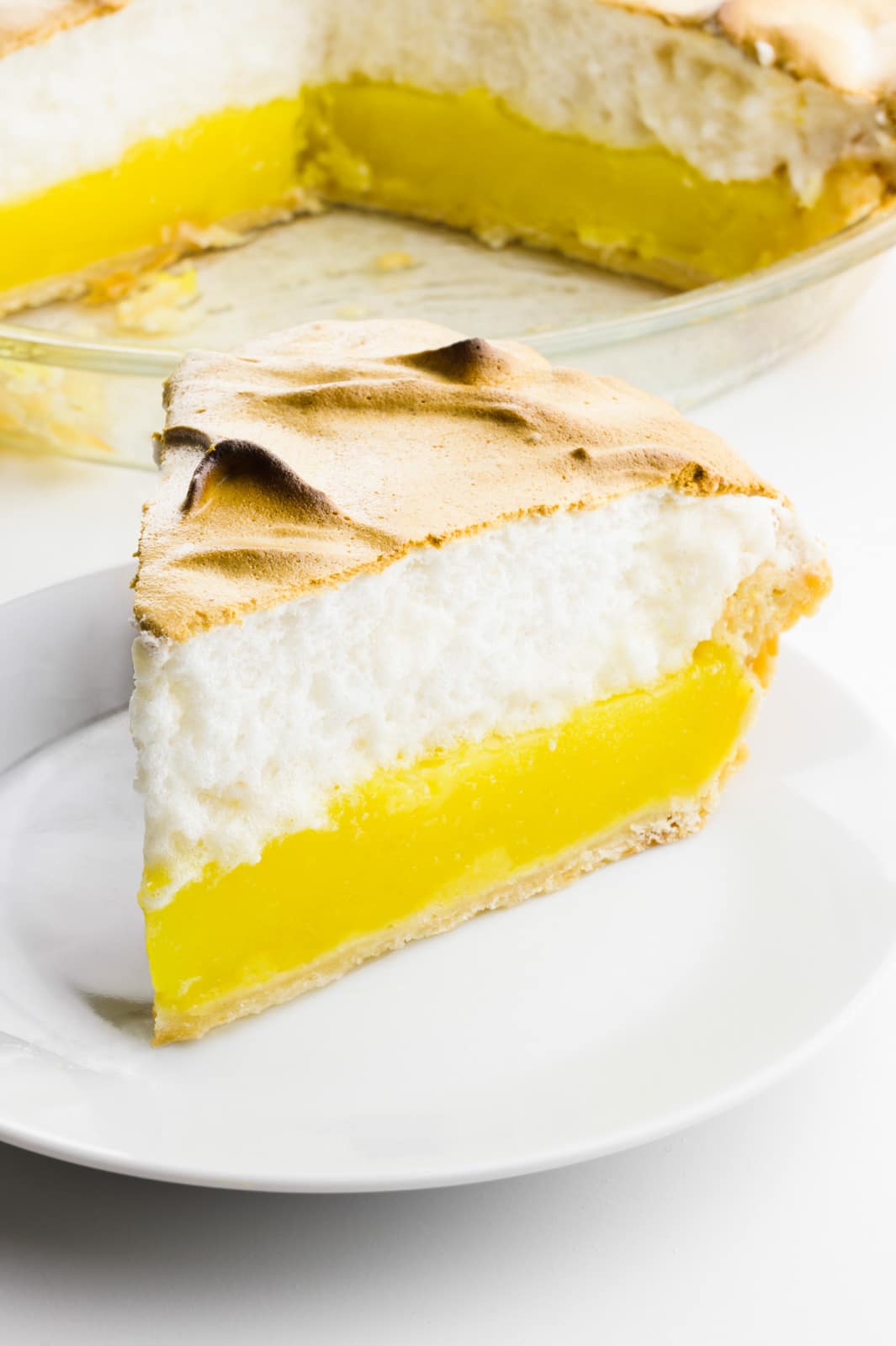 A slice of lemon meringue pie on a plate sits in front of the rest of the pie.