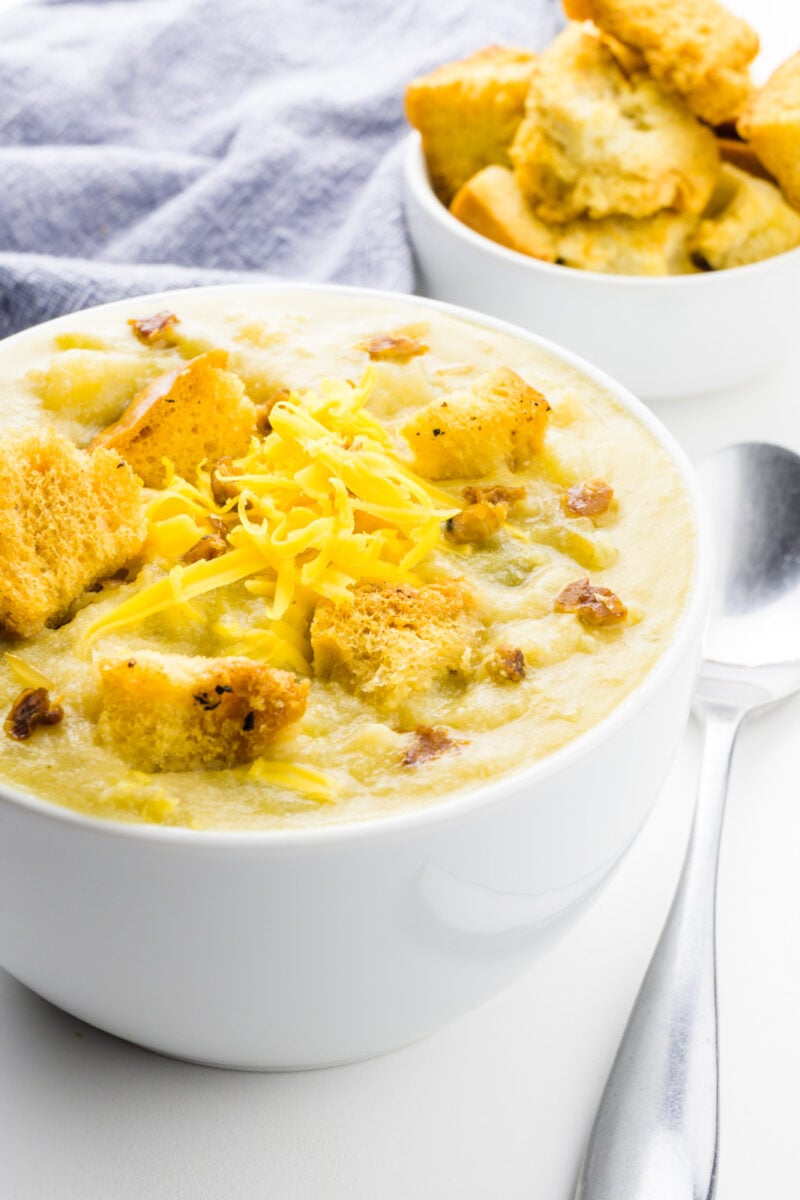 A bowl of vegan potato soup has croups, cheese shreds, and veggie bacon bits on top. A bowl of croutons and a kitchen towel is behind it.