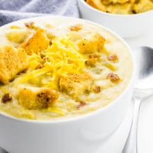 A bowl of vegan potato soup has croups, cheese shreds, and veggie bacon bits on top. A bowl of croutons and a kitchen towel is behind it.