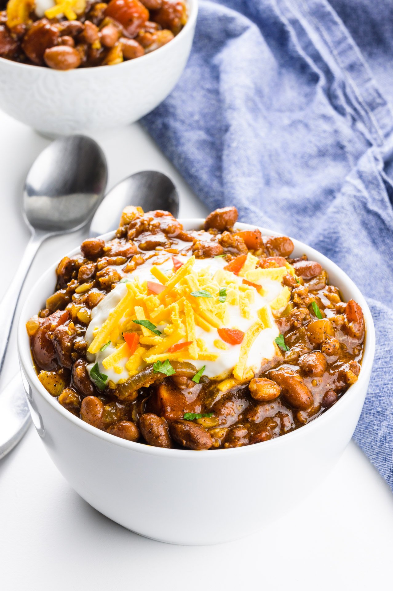 Two spoons sit between two bowls of chili, both topped with vegan sour cream and other toppings. A blue kitchen towel is beside it.