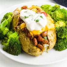 A loaded vegan baked potato sits on a plate with steamed broccoli all around it.