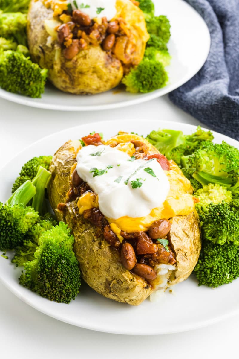 A vegan baked potato sits on a plate with steamed broccoli and loaded toppings. Another plate with a potato is behind it.