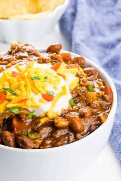A bowl of vegan chili is topped with sour cream, and vegan cheddar shreds. A bowl of tortilla chips and a blue kitchen towel is behind it.