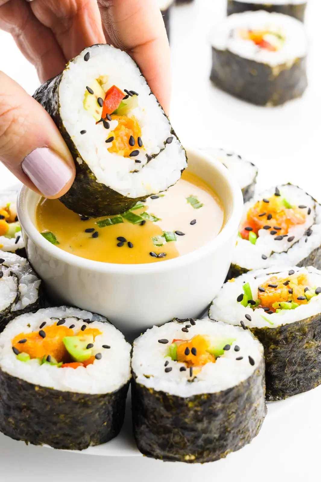 A hand holds vegan sushi, dipping it in a bowl with sauce. There's more sushi rolls around the bowl.