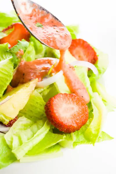 A spoon holds strawberry vinaigrette, hovering over a green salad with red onions and chopped strawberries.