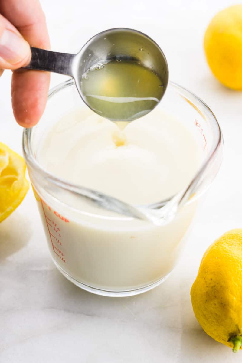 A hand holds a measuring spoon and is pouring lemon juice into plant-based milk in a glass measuring cup.
