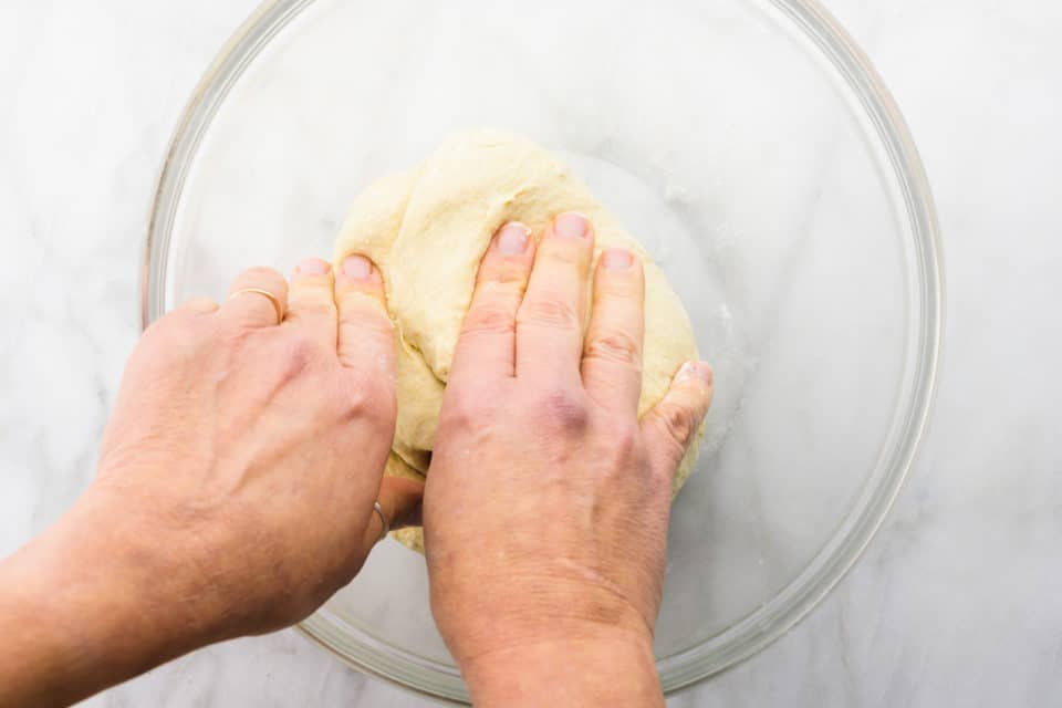 Two hands reach into a glass bowl, kneading vegan pizza dough.