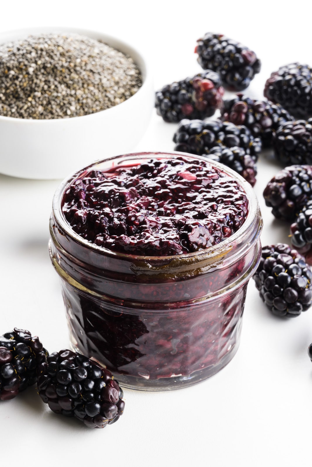 A small jar holds blackberry jam with fresh berries around it and a bowl of chia seeds behind it.