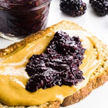 A slice of toast has peanut butter and chia jam on top. There's a jar with more jam behind it and fresh blackberries too.