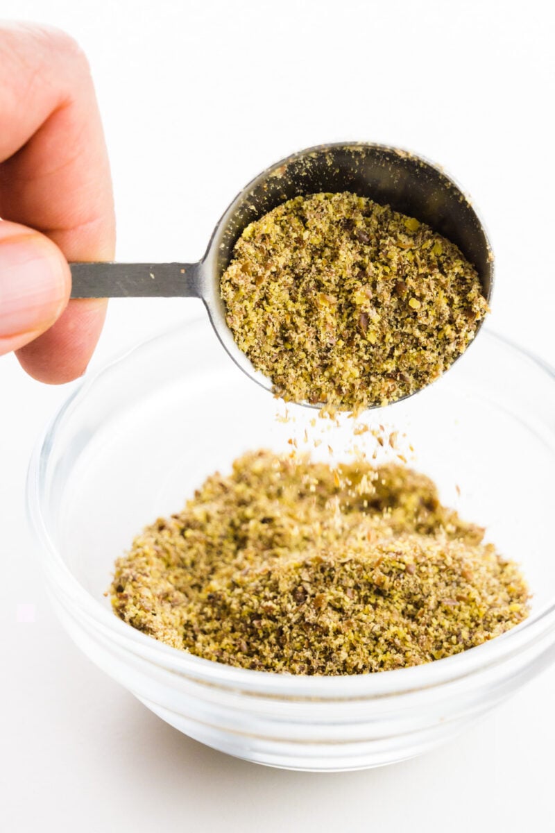 A hand holds a measuring spoon, pouring ground flax seeds into a small bowl.