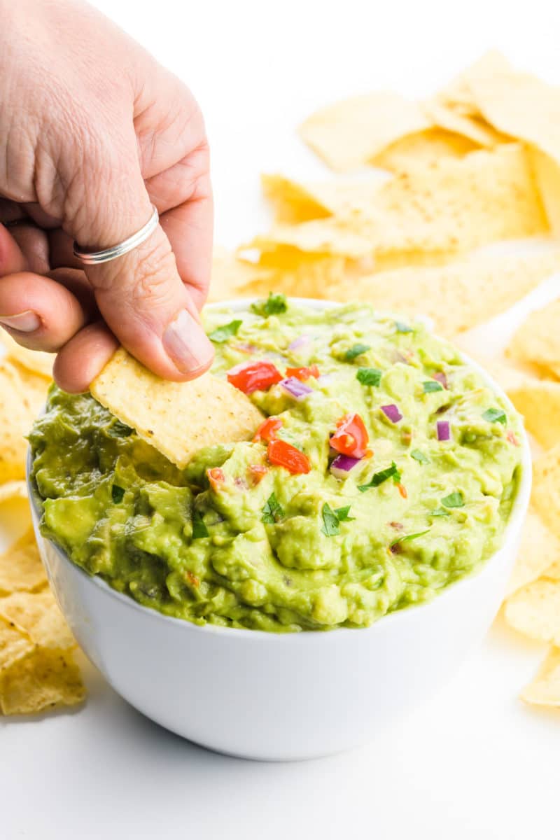 A hand holds a tortilla chip dipping it into a bowl of guacamole. There are tortilla chips behind it.
