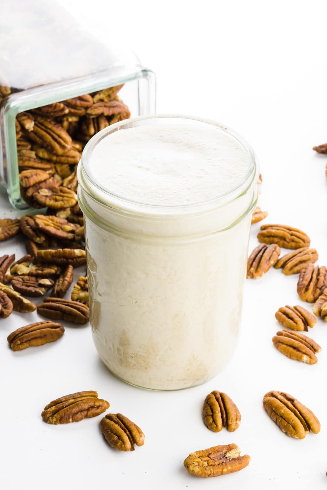 A mason jar holds milk. There's a glass storage container in the background with pecans coming out of it.