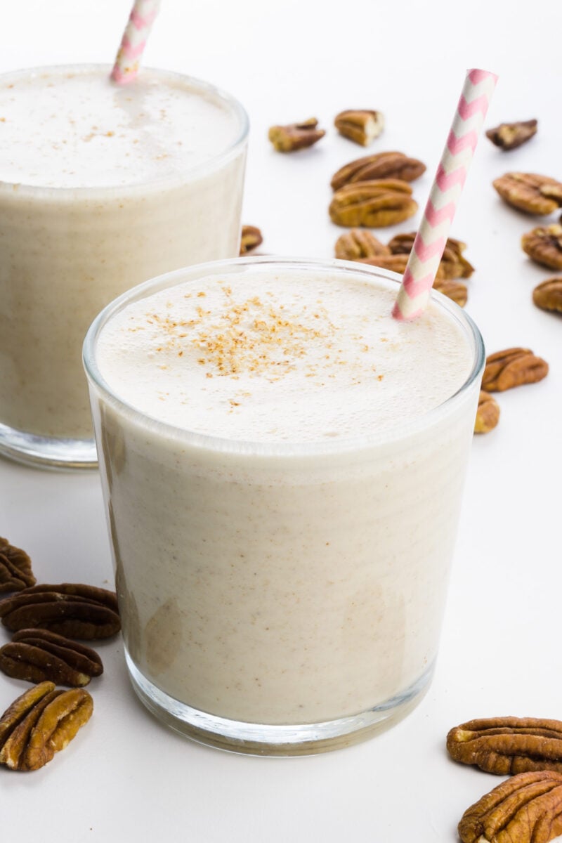 Two glasses hold pecan milk with paper straws. There are pecans all around them.