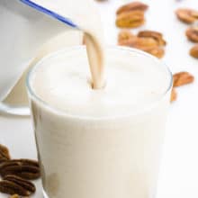 A pitcher pours a creamy milk into a small glass. There are pecans around it.