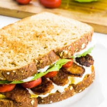 A toasted sandwich sits on a plate. It has chopped tomatoes, tempeh bacon, lettuce, and mayo in it. Behind it is a cutting board with cherry tomatoes and lettuce.