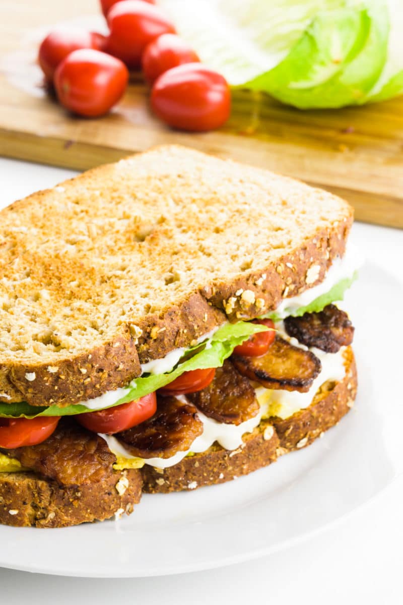 A toasted sandwich sits on a plate. It has chopped tomatoes, tempeh bacon, lettuce, and mayo in it. Behind it is a cutting board with cherry tomatoes and lettuce.