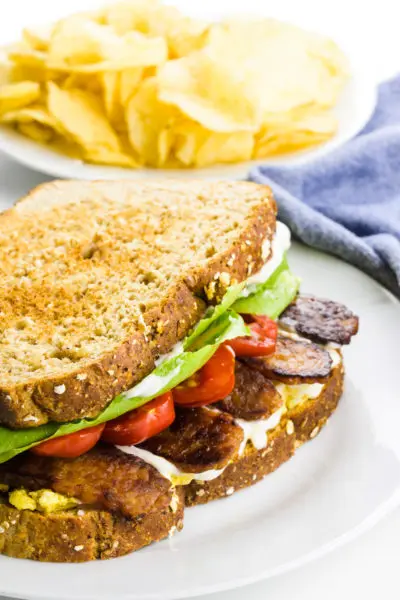 A sandwich has layers of ingredients, including tempeh bacon, tomatoes, lettuce and mayo. There's a plate of potato chips next to a blue kitchen towel in the background.