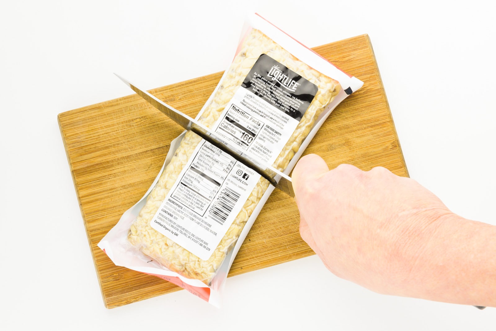 A hand holds a knife, cutting a package of tempeh in half as it sits on a cutting board.