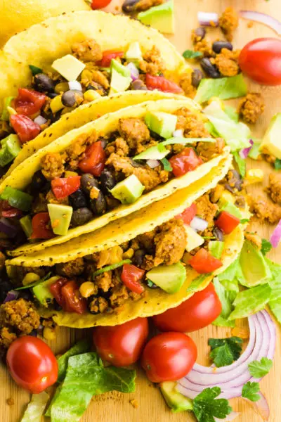 Three tacos are next to each other with ingredients all around, like cherry tomatoes, greens, and more.