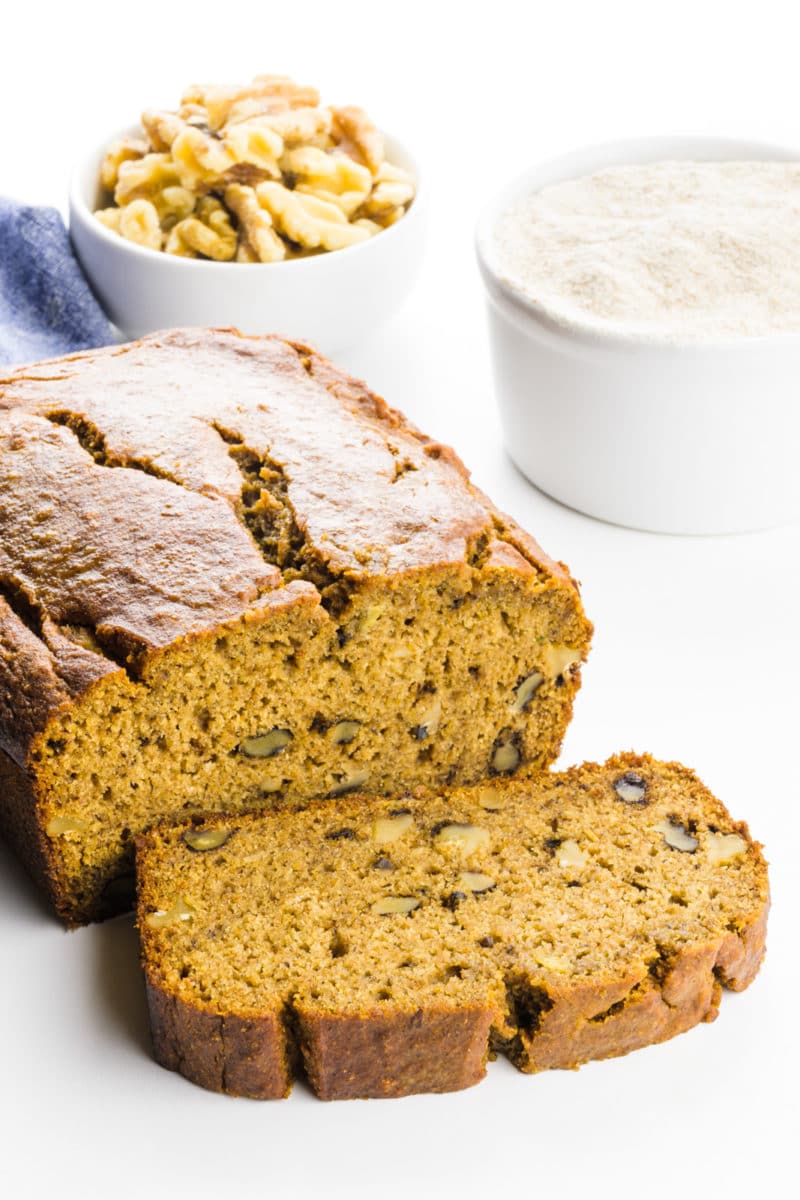 A slice of banana bread sits in front of the rest of the loaf. There's a measuring cup with whole wheat flour and a bowl with walnuts behind it.