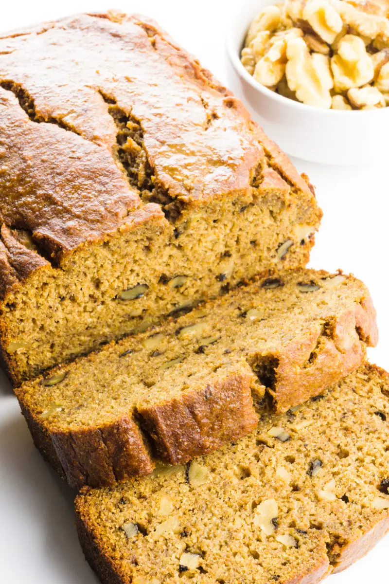 A loaf of whole wheat banana bread has several slices cut out. There's a small bowl of walnuts behind it.