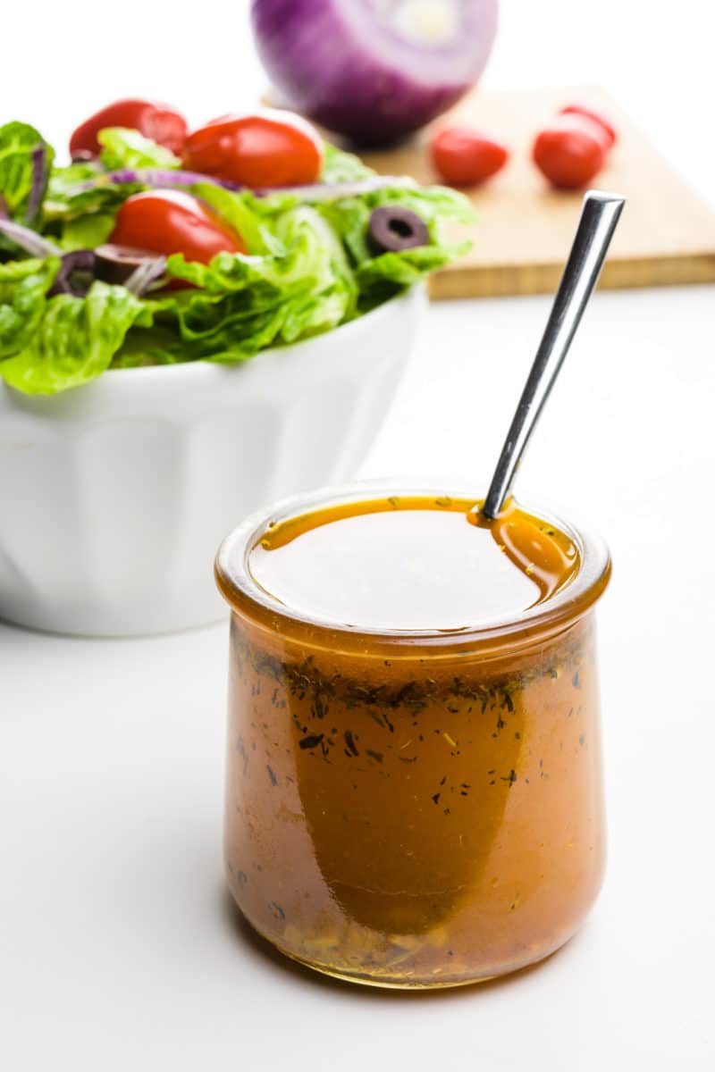 A jar of Italian vinaigrette has a spoon in it. It's sitting in front of a salad and a cutting board with more salad ingredients.