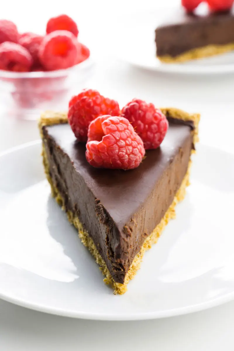 A slice of chocolate pie on a plate has a bowl of raspberries and another slice of pie behind it.