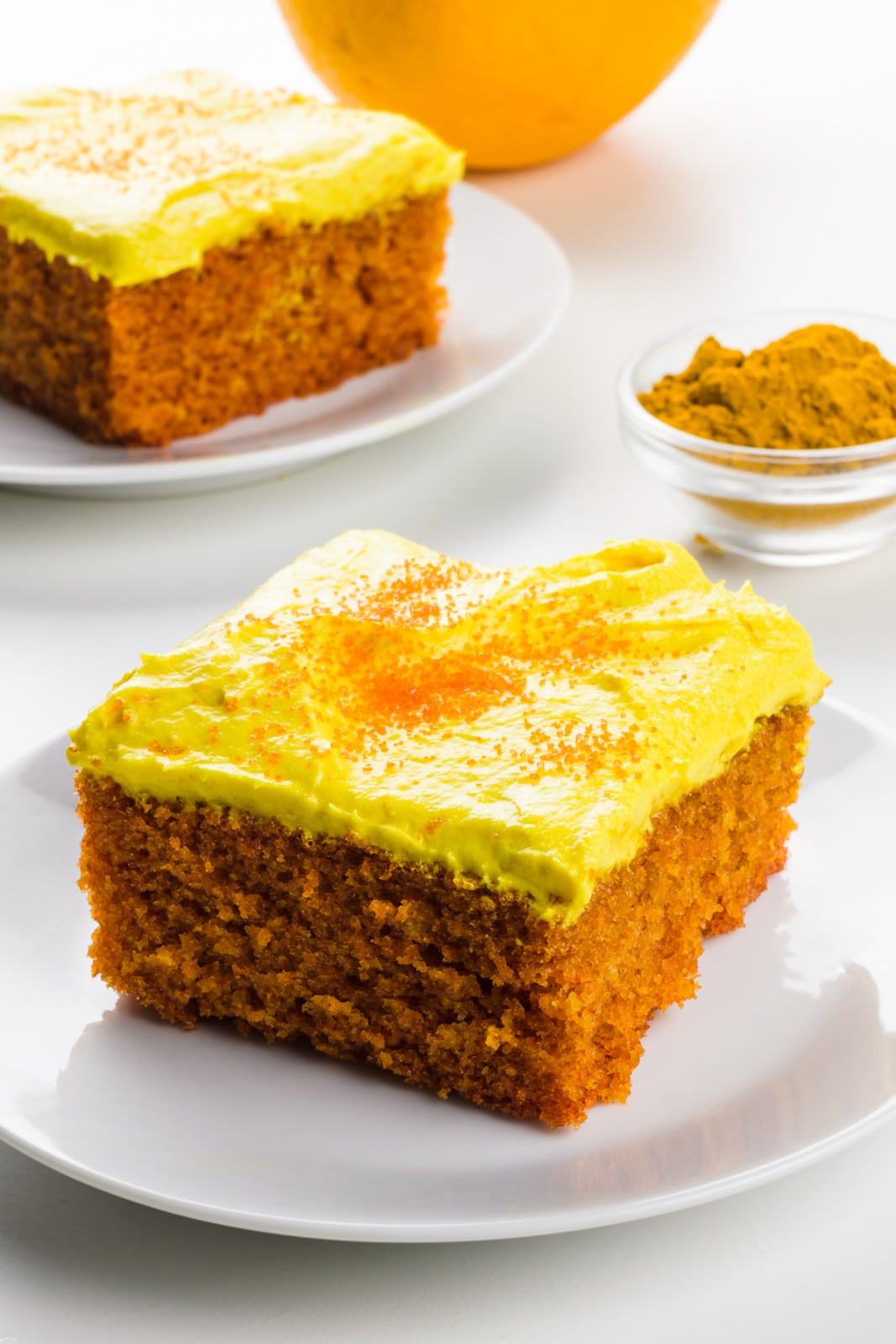 A slice of turmeric cake on a plate with another slice behind it. There's an orange and a bowl of turmeric beside it.