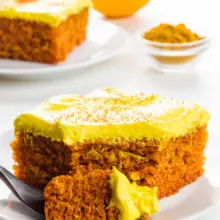 A slice of turmeric cake sits on a plate with a bite sitting on a fork. An orange and a small bowl of turmeric is behind it.