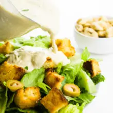 A serving dish full of vegan caesar dressing is being poured over a salad. There's a bowl of cashews in the background.