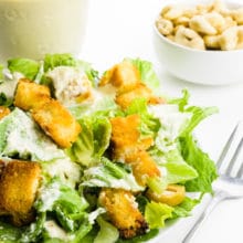 A vegan caesar salad is topped with croutons, olives, and dressing. There's a fork, more dressing, and cashews next to it.