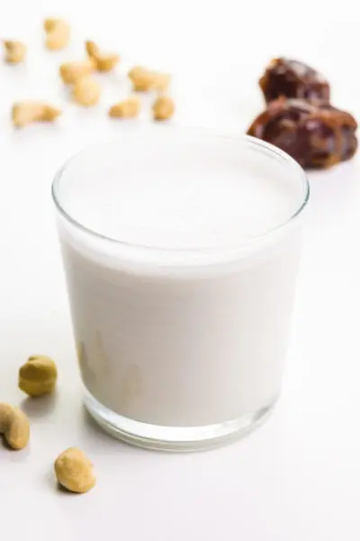A glass of milk has cashews around it and some dates behind it.