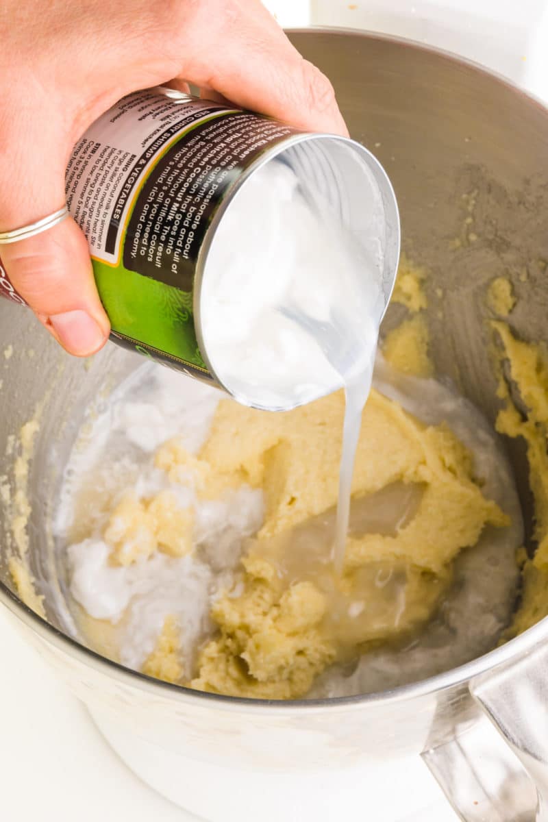 A hand holds a can of coconut milk pouring it into a mixing bowl along with a butter mixture. 
