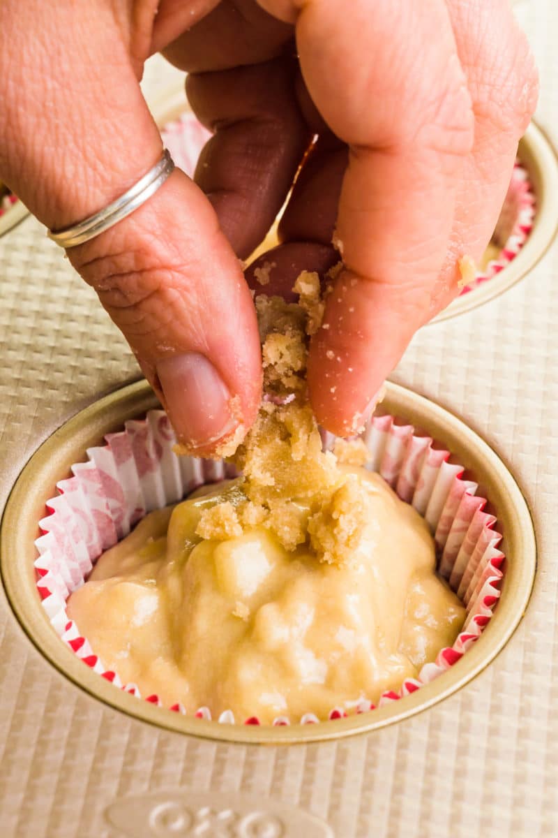 A hand is placing crumble topping on top of muffin batter in a muffin compartment lined with paper.