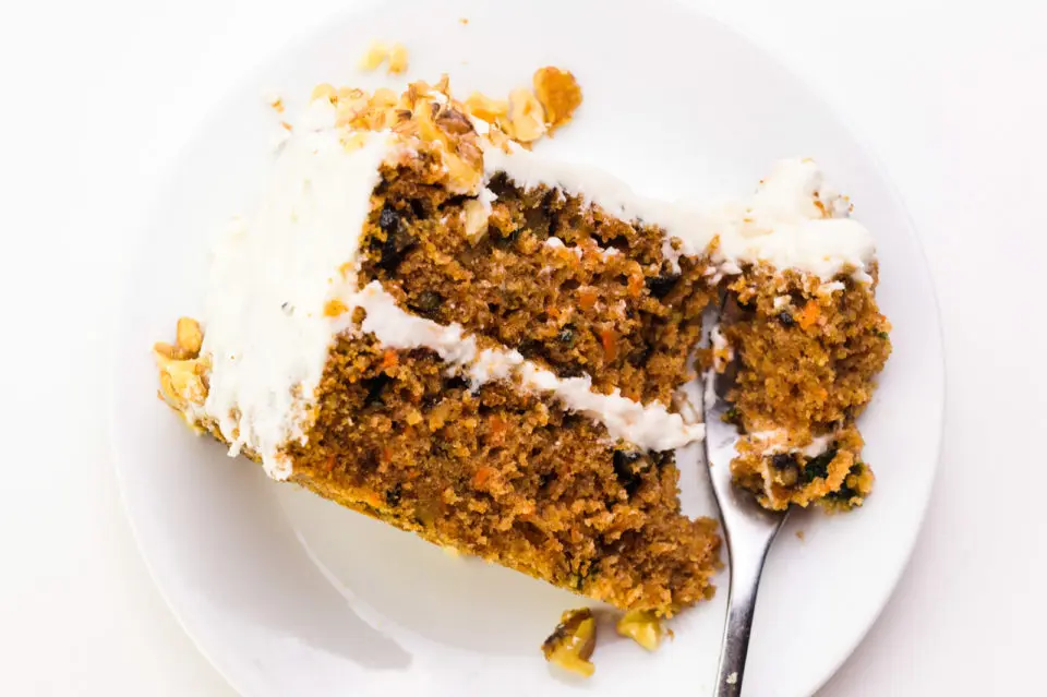 Looking down on a plate with a slice of carrot cake on it. A fork has cut into the cake and is sitting at the front of the plate.