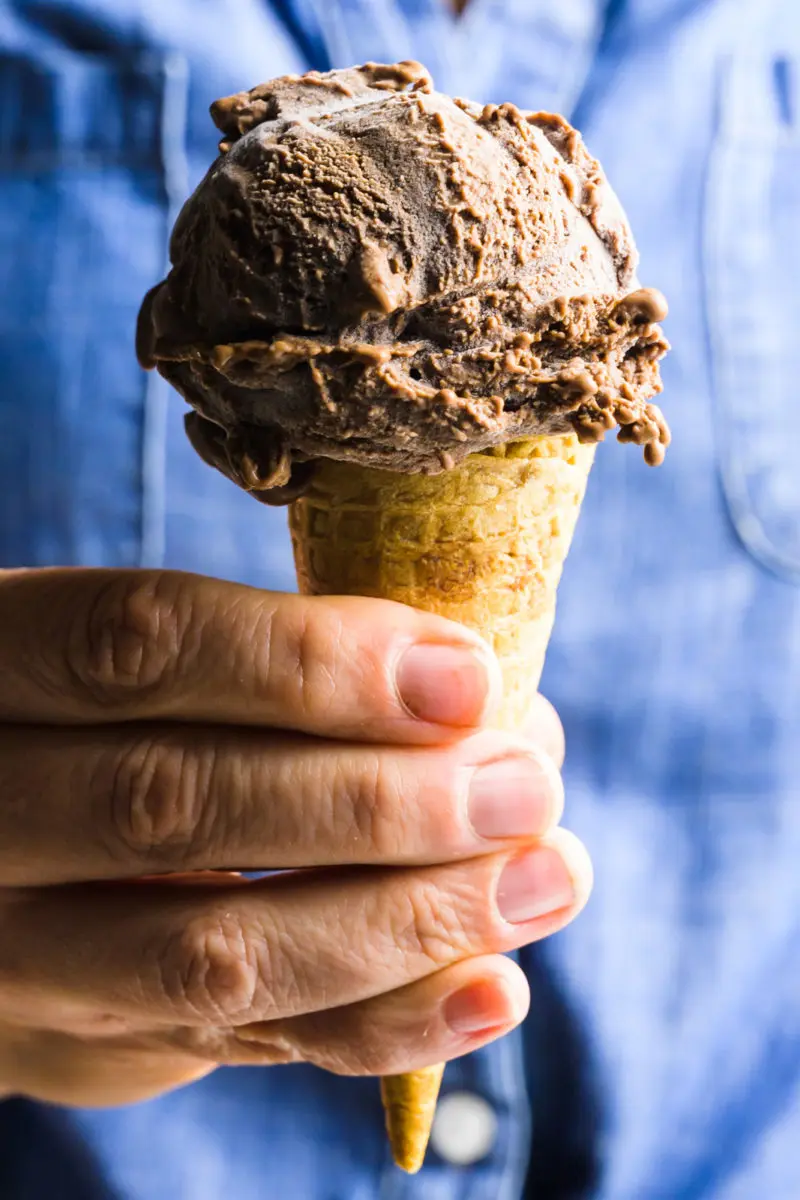 A hand holds a cone full of a large scoop of vegan chocolate ice cream. The person holding it has on a blue top.