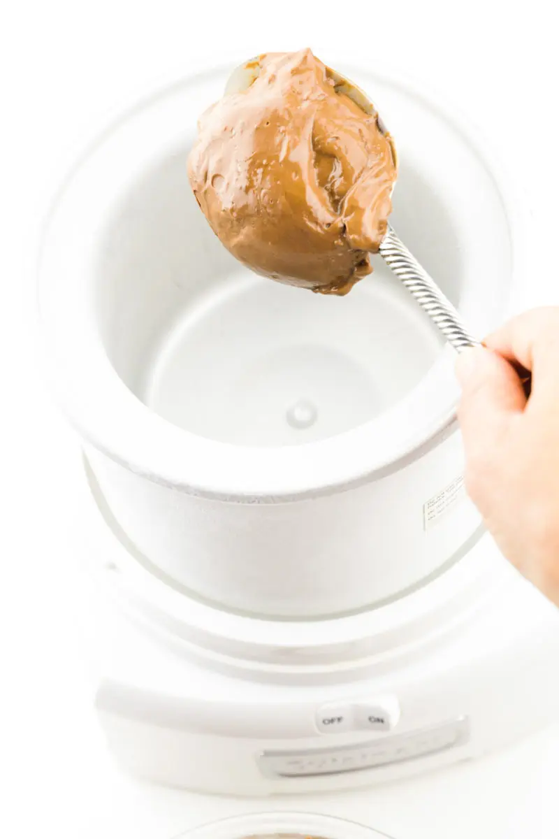 A hand holds a spoon dropping a chocolate mixture into a chilled ice cream maker bowl.
