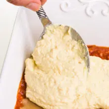 A hand holds a spoon, spreading vegan ricotta over lasagna noodles and sauce in a baking dish.