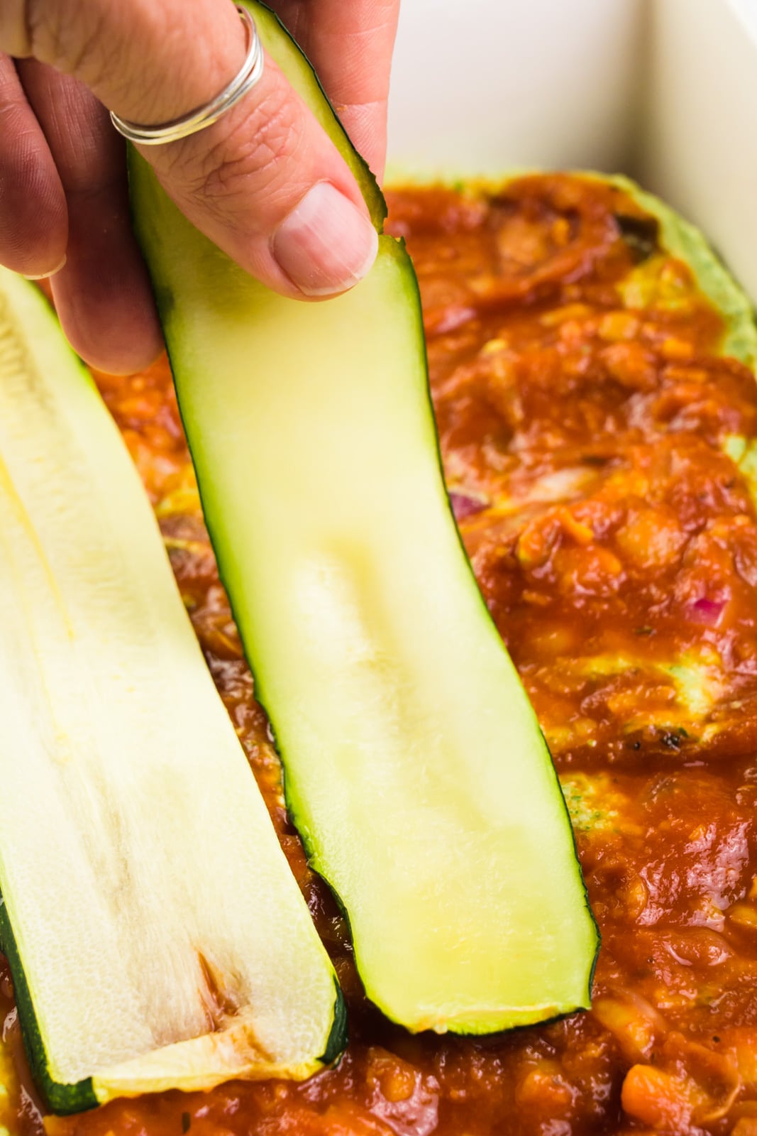 A hand is placing a zucchini slice over red sauce in a baking dish.