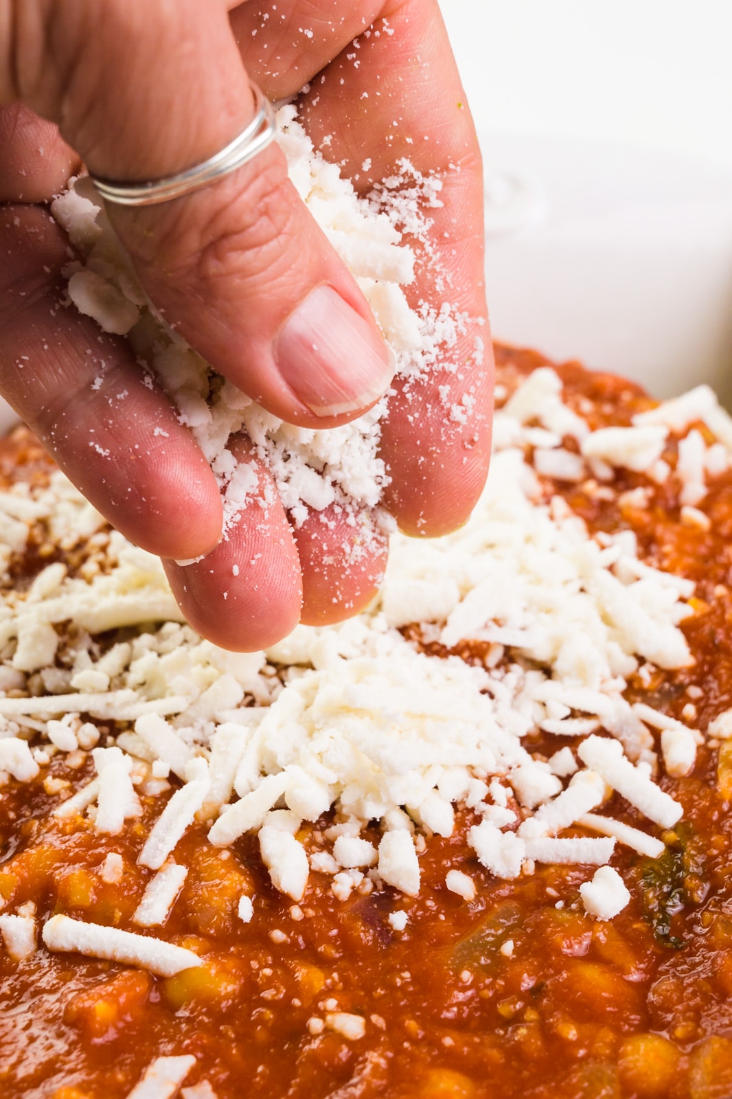A hand is distributing mozzarella cheese over red sauce in a baking dish.
