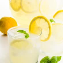 Two glasses of lemonade have fresh lemon slices on the glasses. There's a lemon behind it, and basil sprigs in front of it. There's a pitcher of more of the beverage behind it.