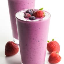 Two glasses are full of bright pink mixed berry smoothie mixtures. Both have berries on top and strawberries on the white counter beside them.