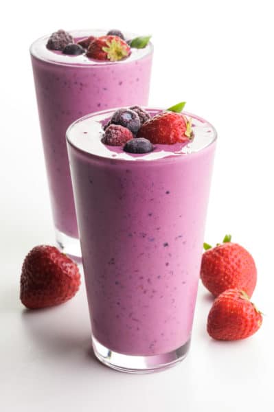 Two glasses are full of bright pink mixed berry smoothie mixtures. Both have berries on top and strawberries on the white counter beside them.