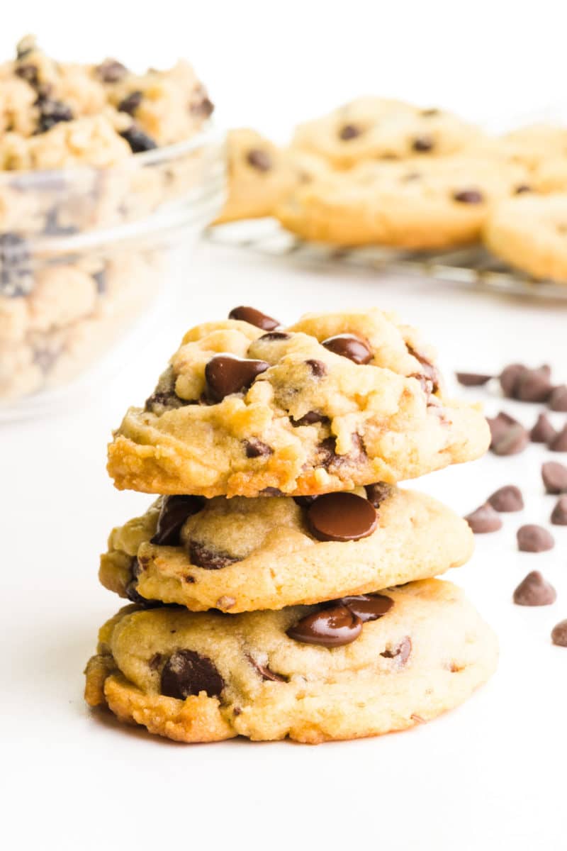 A stack of vegan chocolate chip cookies sit in front of a bowl of cookie dough, chocolate chips, and more cookies on a wire rack.