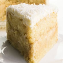A slice of vegan coconut cake on a plate with the rest of the cake behind it.