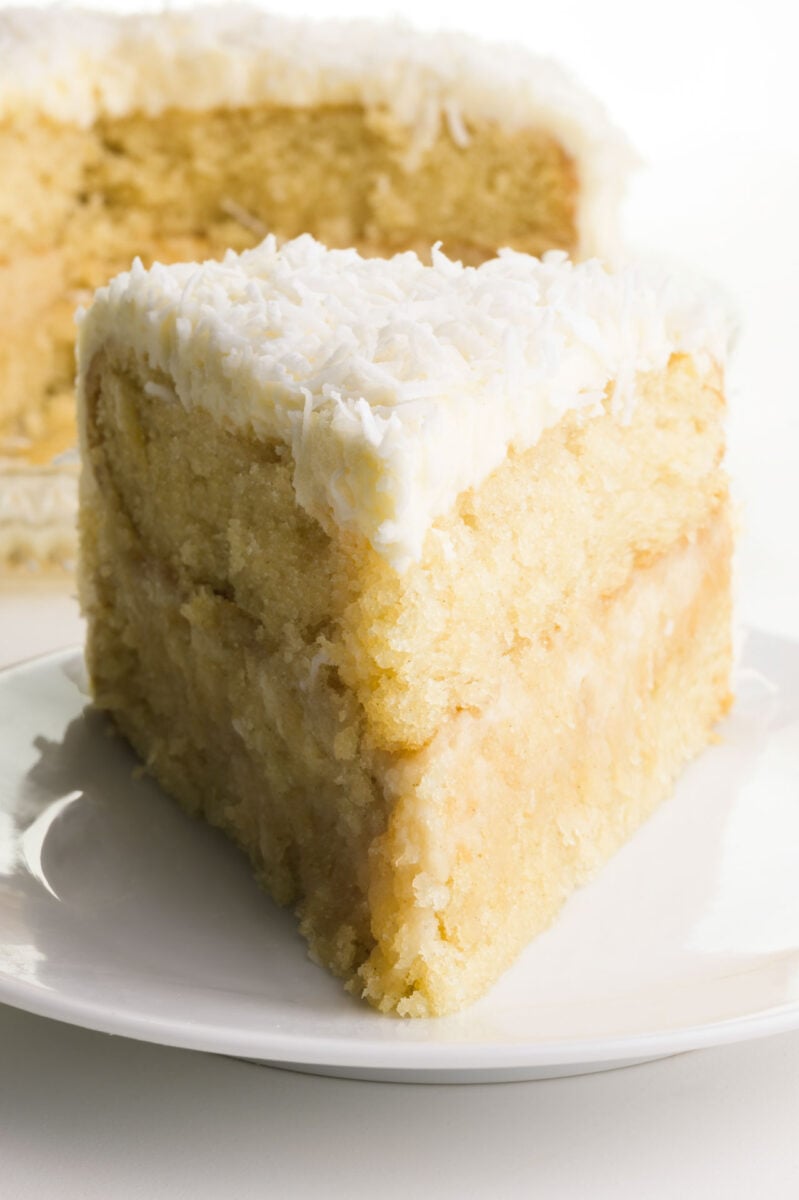 A slice of vegan coconut cake on a plate with the rest of the cake behind it.